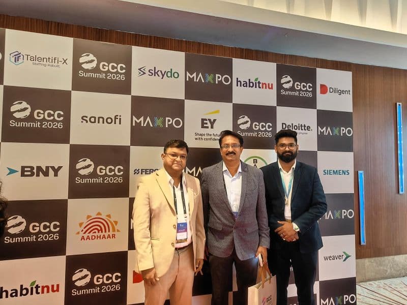 Three members of the TalentiFi-X team in business attire at the GCC Summit 2026 sponsor backdrop in Bengaluru.