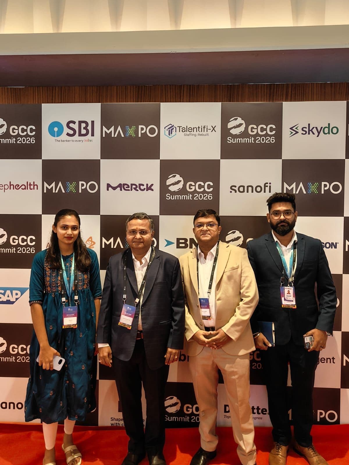 Four TalentiFi-X team members wearing exhibitor badges at the GCC Summit 2026 sponsor backdrop.